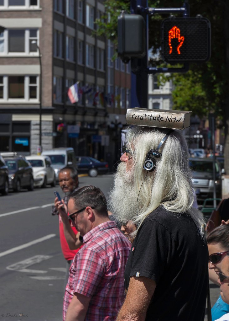 grey haired man with headphones and Gratitude Now book on his head walks past a Do Not Cross hand sign